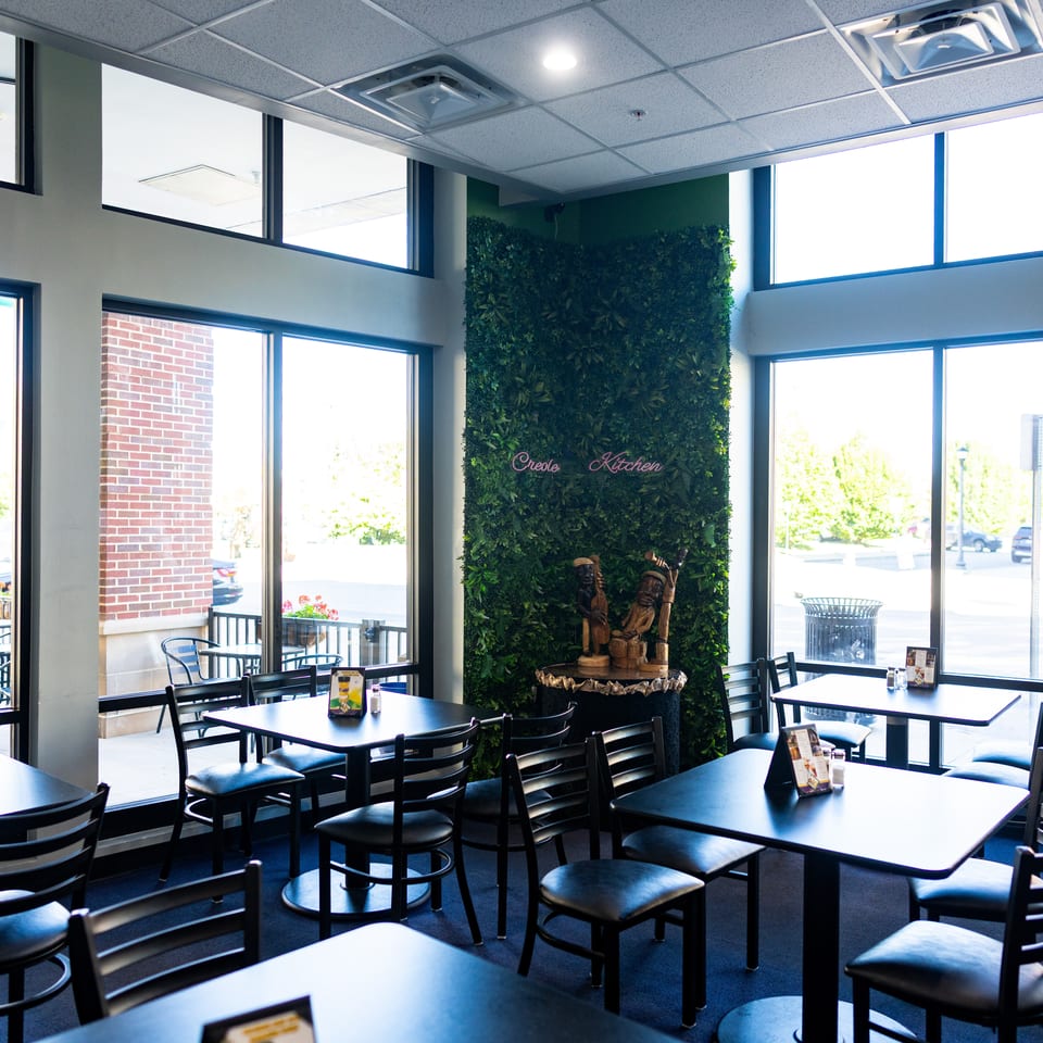 Bright Creole Kitchen dining room with black tables, chairs, large windows, and a leafy wall featuring a fountain and sculptures.