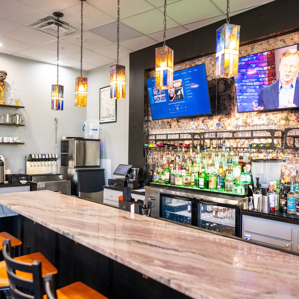 Modern restaurant bar with colorful pendant lights, liquor bottles, TVs, counter seating, and coffee equipment.