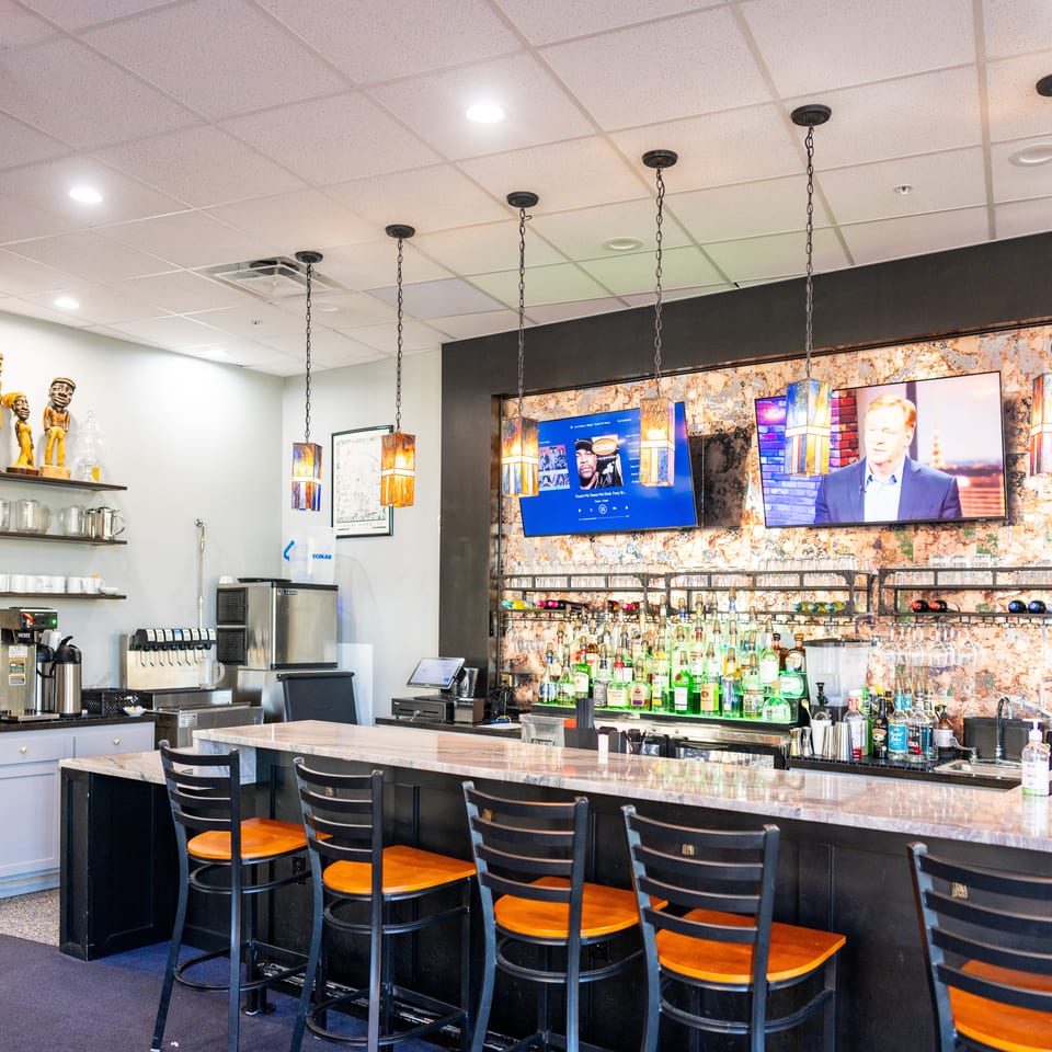 Restaurant bar with colorful pendant lights, liquor bottles, television screens, and rows of black chairs with orange seats.