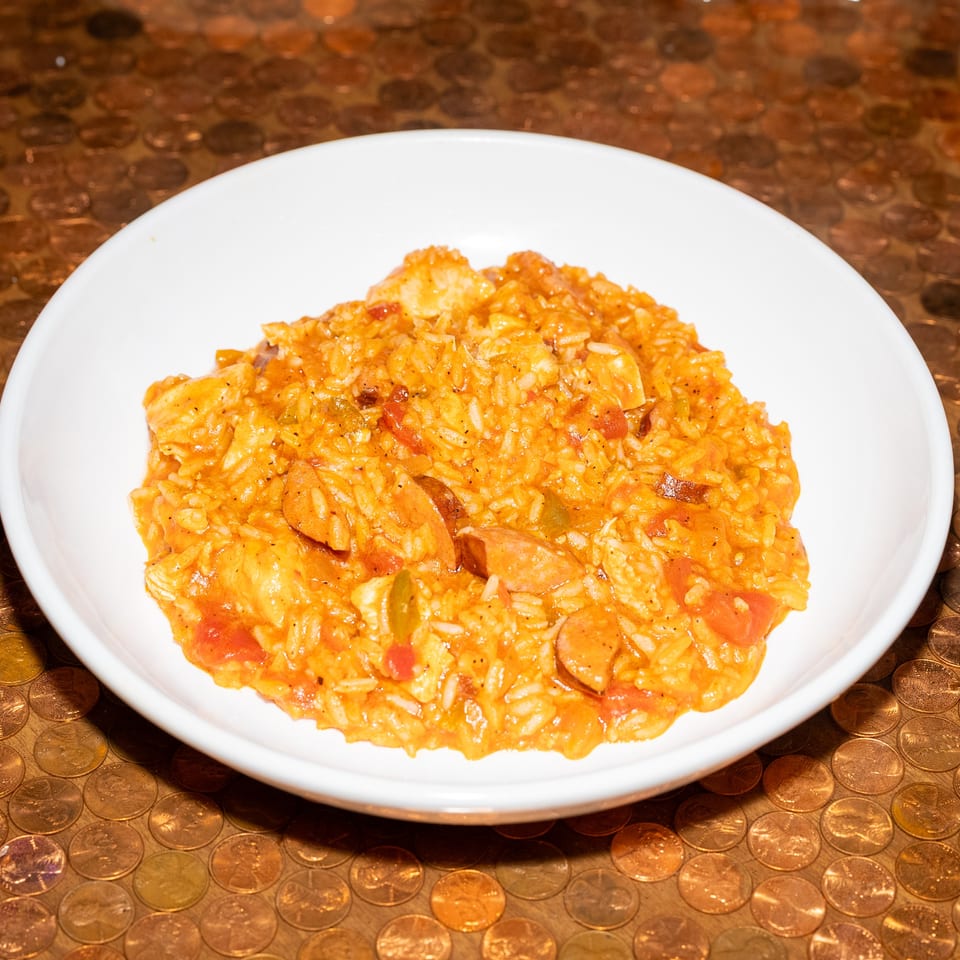White bowl of orange rice mixed with sliced sausage, vegetables, and tomatoes on a coin-patterned tabletop.