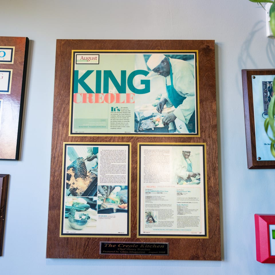 Framed Ohio Magazine feature about The Creole Kitchen shows Chef Henry Butcher preparing food, surrounded by restaurant memorabilia and greenery.