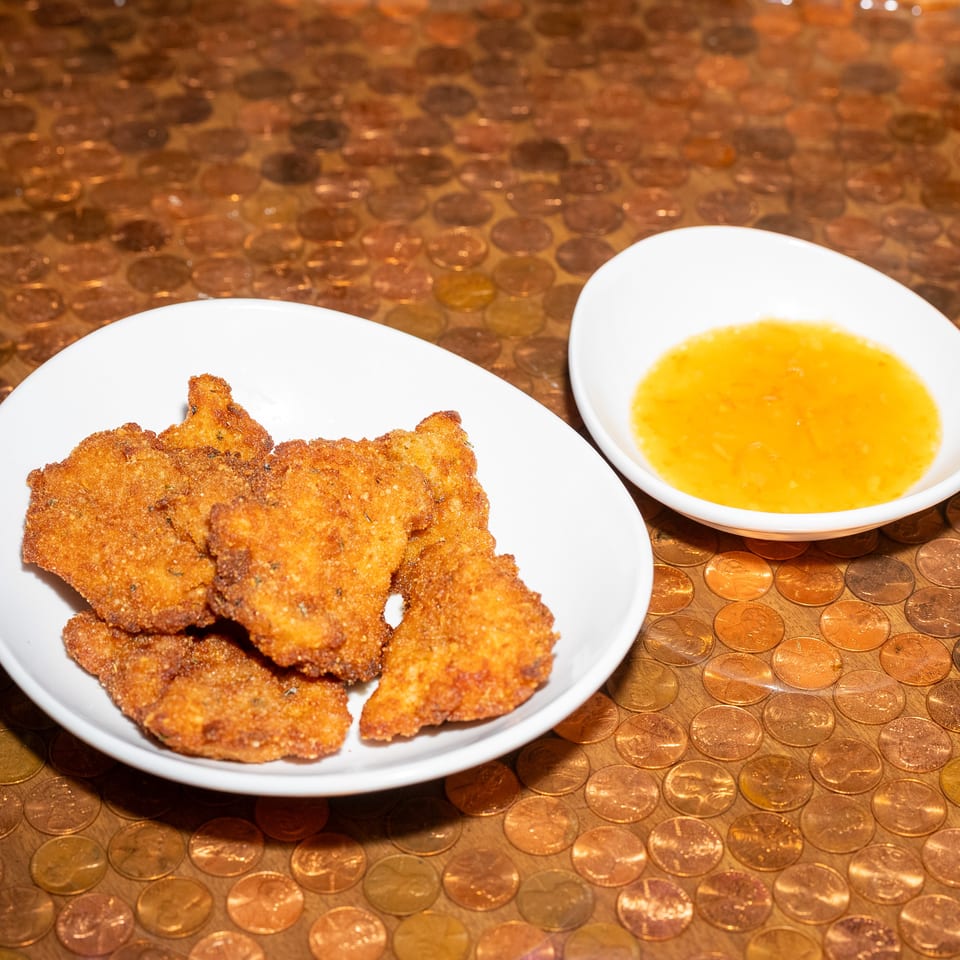 Crispy breaded appetizers on a white plate with orange dipping sauce, set on a tabletop covered in copper-colored coins.