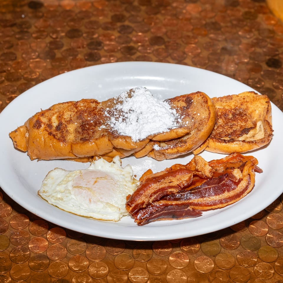 Plate of powdered-sugar French toast with fried eggs and crispy bacon on a copper coin-patterned table.