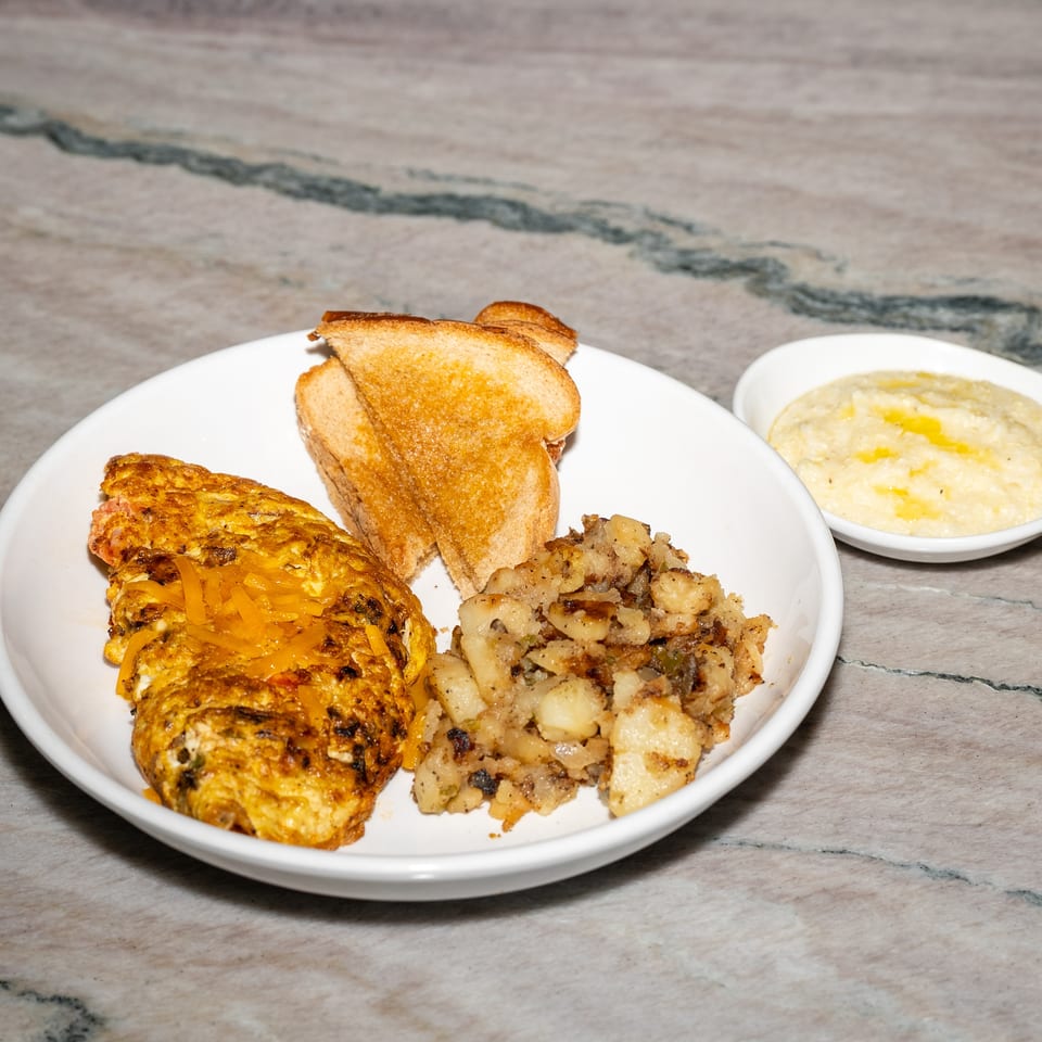 Breakfast plate with cheesy omelet, home fries, toast, and a side bowl of creamy grits on a marble countertop.