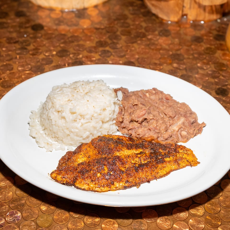 White plate of blackened fish, white rice, and refried beans on a table covered with copper-colored coins.