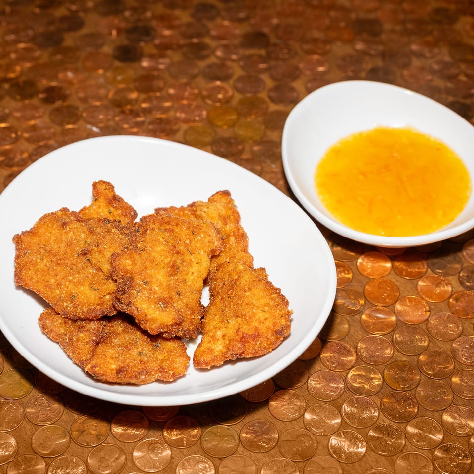 Golden breaded fried cutlets on a white plate with a bowl of orange dipping sauce on a coin-covered tabletop.