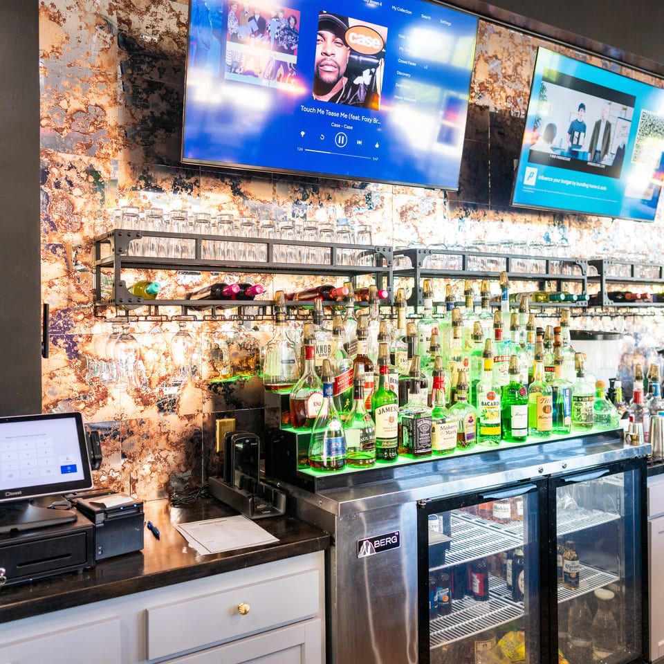 Restaurant bar stocked with liquor bottles and glasses, illuminated beneath shelves and televisions displaying music and advertisements.