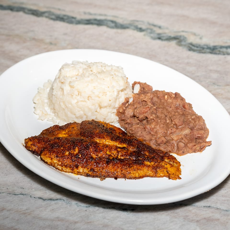 Plate of seasoned grilled fish with white rice and refried beans on a marble tabletop.