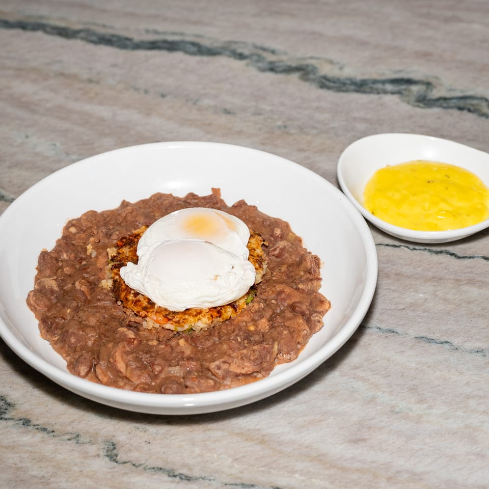 Refried beans topped with a crispy rice-and-vegetable cake and two poached eggs, served with yellow sauce on a marble table.