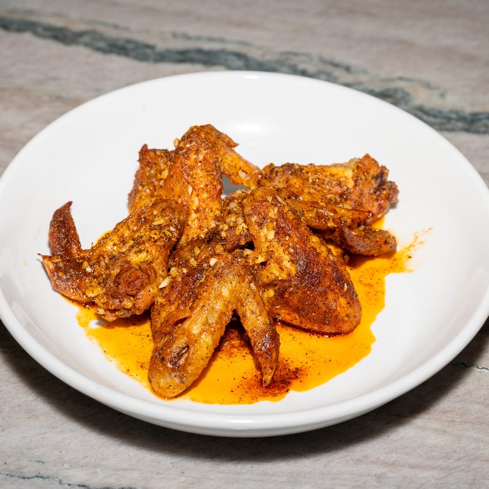 Spiced, crispy chicken wings piled on a white plate with orange chili oil, resting on a marble tabletop.