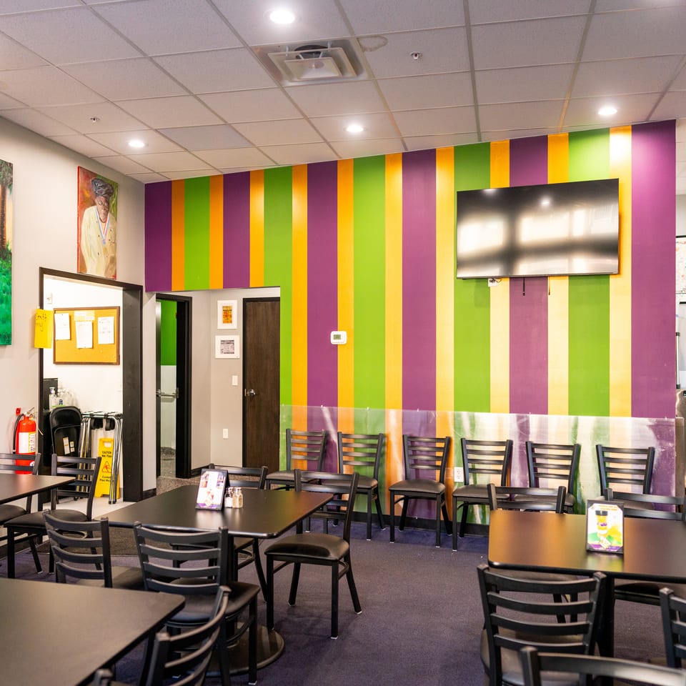 Restaurant dining area with black tables and chairs, colorful striped wall, mounted television, and cultural artwork.