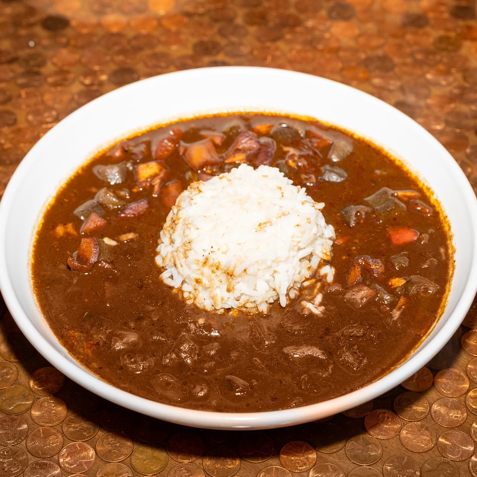 White rice surrounded by dark brown stew with vegetables and meat in a bowl on a copper coin-covered table.