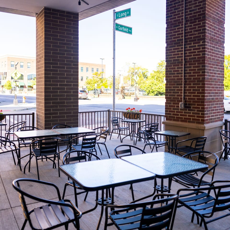 Outdoor restaurant patio with black tables and chairs, brick columns, flower planters, and E Long Street and N Garfield Avenue signs.