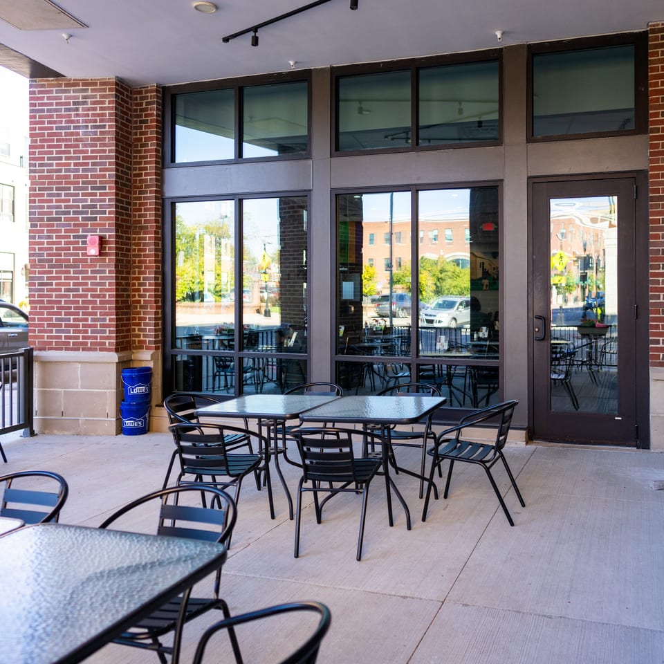 Covered restaurant patio with black tables and chairs beside a brick building entrance, overlooking a city street.